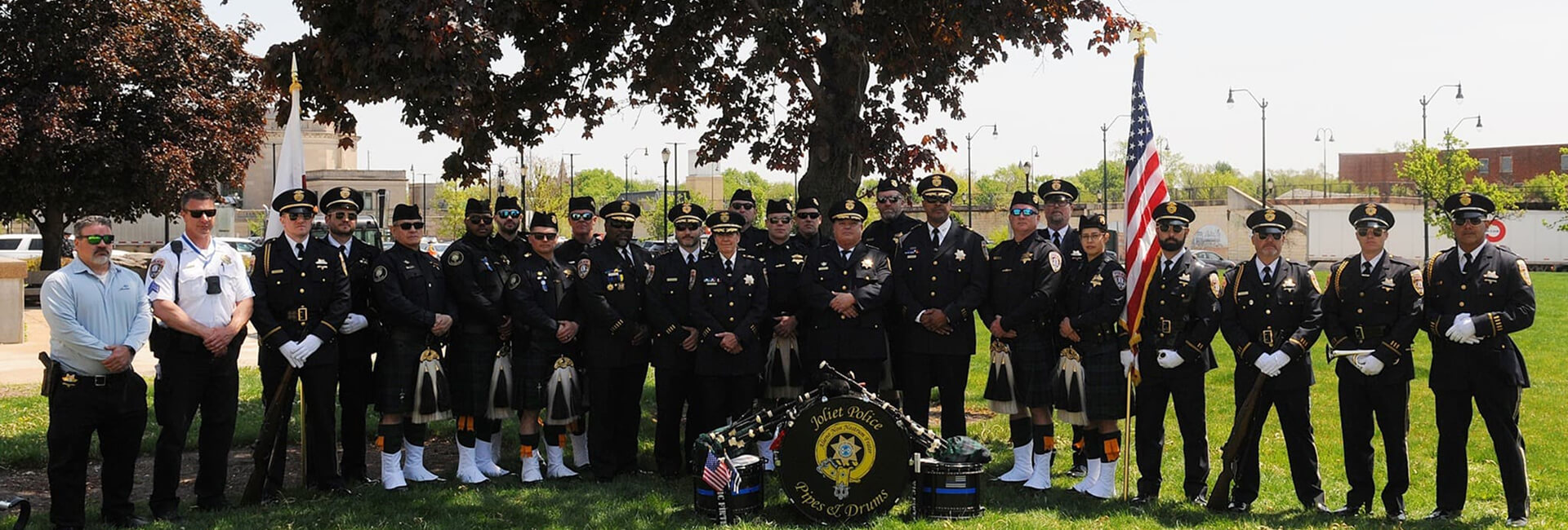 Joliet Police Pipes and Drums members and police officers pose beneath a tree with the American flag.